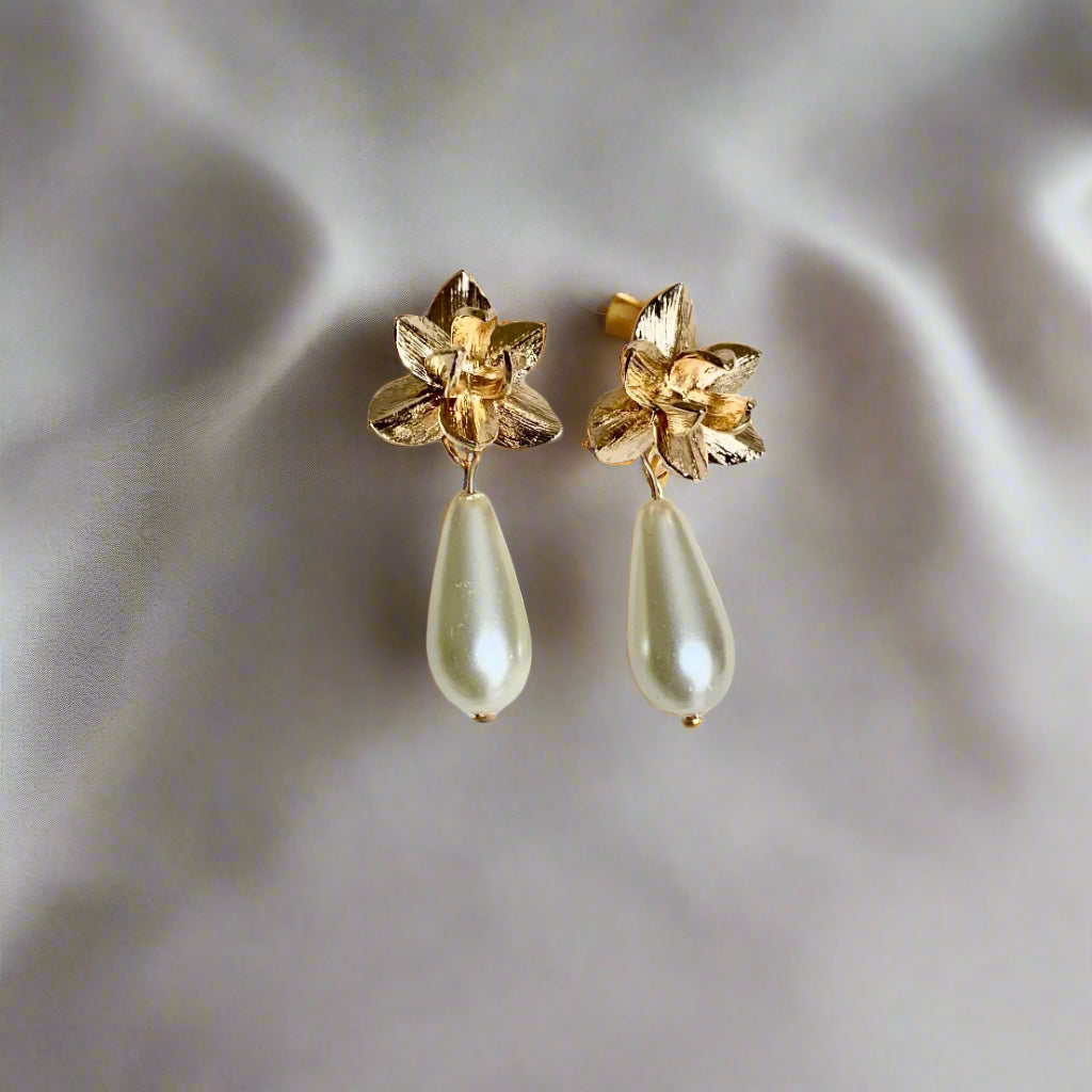 A pair of gold-colored teardrop pearl earrings with floral designs on the top.
