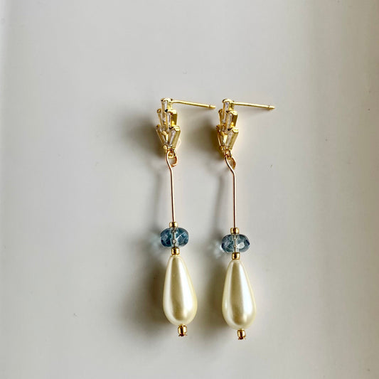 A pair of teardrop pearl and blue crystal earrings dangle from gold-colored metal settings against a white background.